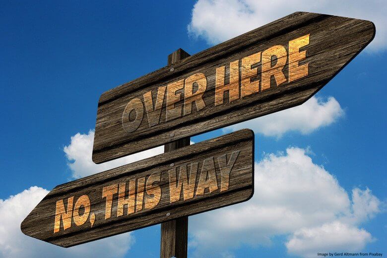 Wooden arrow pointing left with "No, this way" and right with "over here" in front of blue sky with white clouds